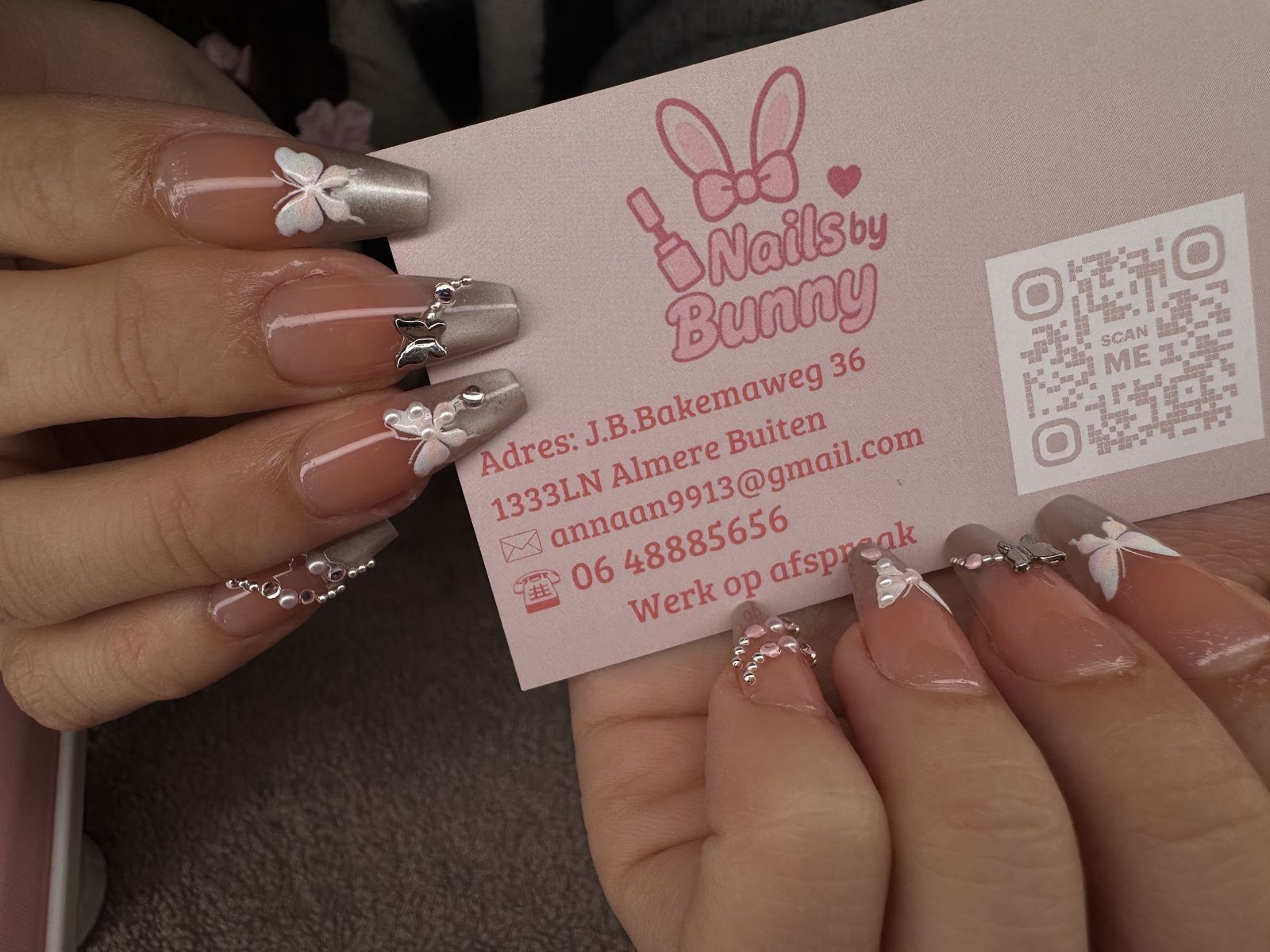 Taupe amandelnagels met witte vlinders en zilveren details, met het visitekaartje van Nails by Bunny in de hand
